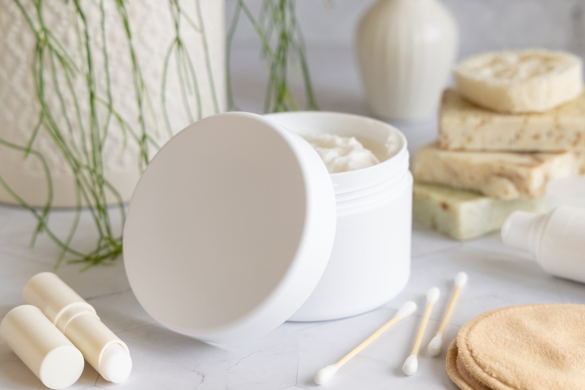 Opened cream jar with white blank lid on marble near green plant close up, cosmetic mockup